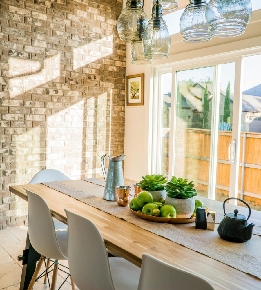 Sunlit dining room with contemporary decor and a rustic brick wall, featuring stylish lighting and a wooden table.